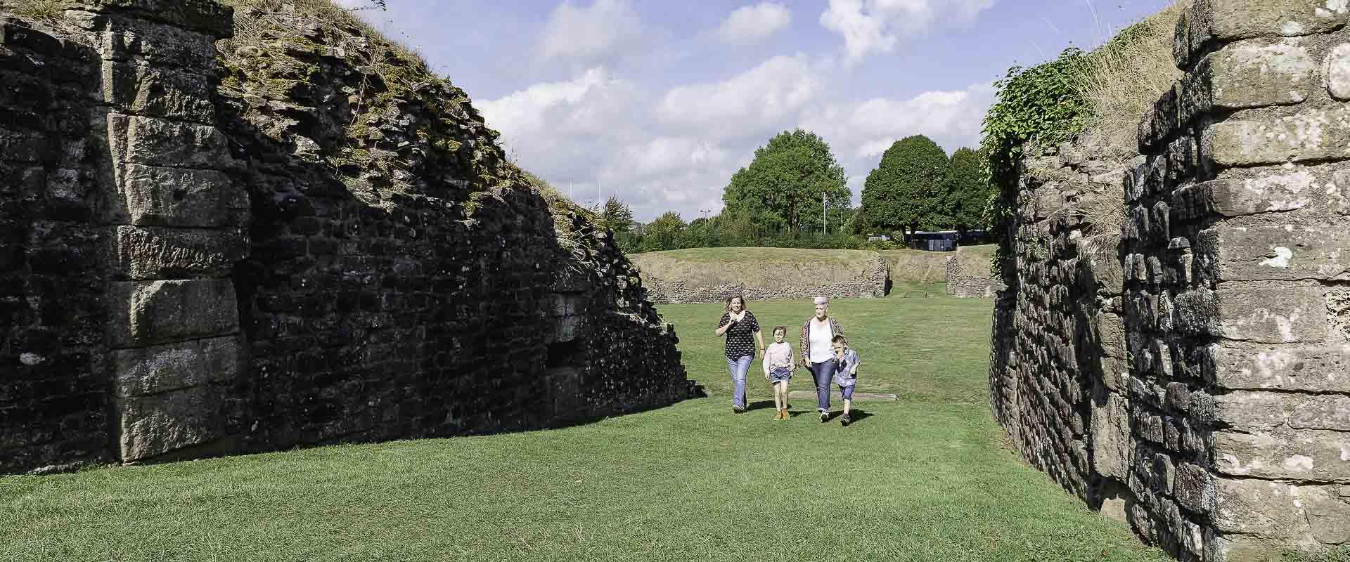 Ymwelwyr ag amffitheatr Caerllion rhwng y sylfeini ar gyfer y stondinau gwylio/Visitors to Caerleon amphitheatre between the foundations for the viewing stands.
